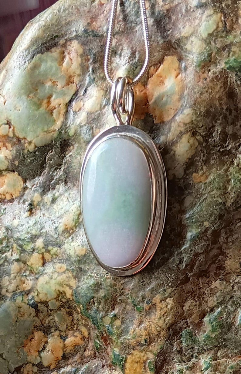 Op de afbeelding: Ovale jade hanger aan een zilveren ketting. De jade steen heeft een lichtgroene en witte kleur. De hanger is gezet in een zilveren bezel. De ketting is een dunne, zilveren slangenketting. De achtergrond is een getextureerde, groen-bruine steen.
