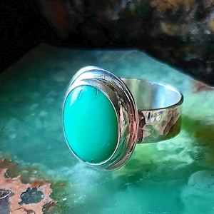 May include: A silver ring with a large, oval, bright green stone. The ring has a hammered texture on the band and a smooth, polished setting around the stone. The ring is displayed on a green surface.