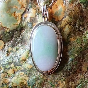 Op de afbeelding: Ovale hangerketting met een lichtgroene en witte steen gezet in een zilverkleurig metalen frame. De hanger hangt aan een delicate zilveren ketting. De achtergrond is een getextureerde, veelkleurige steen.