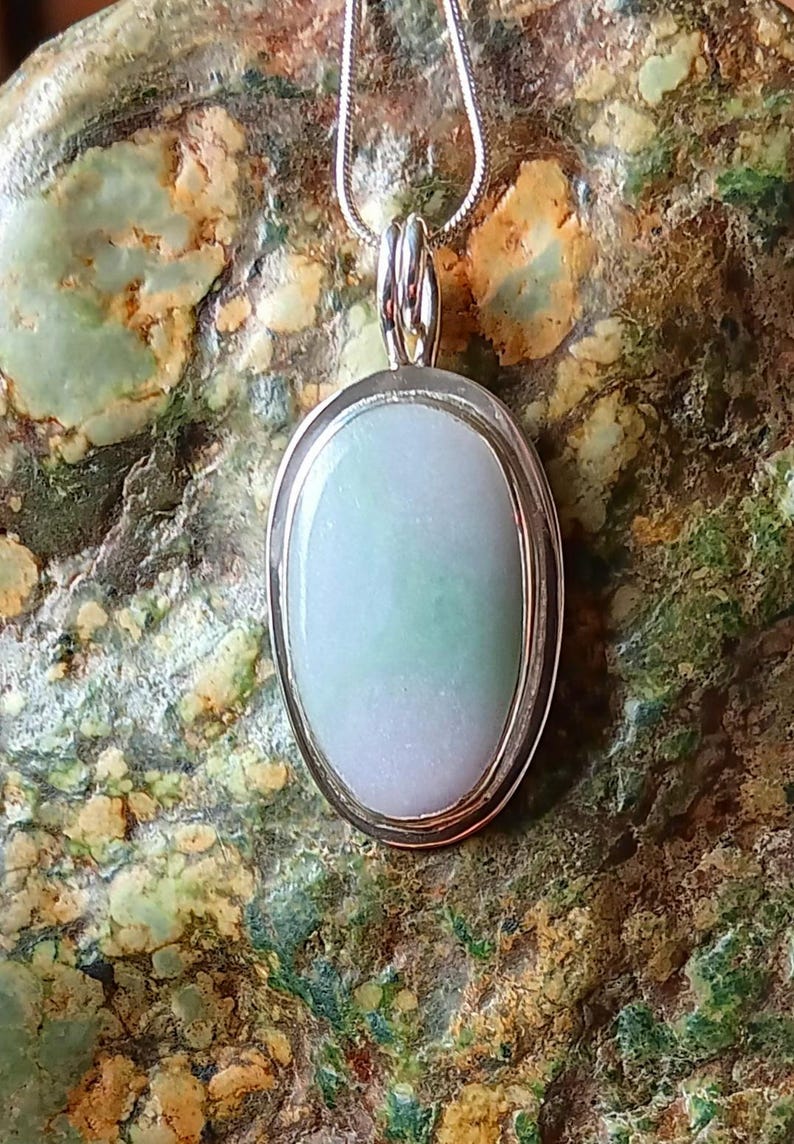 Op de afbeelding: Ovale hangerketting met een lichtgroene en witte steen gezet in een zilverkleurig metalen frame. De hanger hangt aan een delicate zilveren ketting. De achtergrond is een getextureerde, veelkleurige steen.