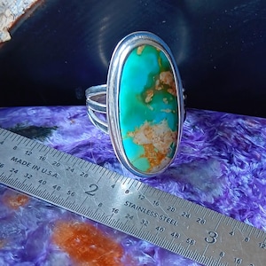 May include: A silver ring featuring a large, oval-shaped turquoise stone with brown matrix inclusions. The ring is positioned next to a stainless steel ruler, showing the stone measures approximately 1.25 inches in length.