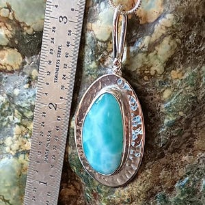 K&ouml;nnte beinhalten: Ovale Anh&auml;nger Halskette mit einem gro&szlig;en, hellblauen Edelstein, ca. 3,8 cm lang, in einem silberfarbenen Metallrahmen gefasst. Der Anh&auml;nger h&auml;ngt an einer zarten Silberkette. Der Edelstein hat ein marmoriertes Aussehen. Der Hintergrund ist eine gesprenkelte gr&uuml;n-braune Oberfl&auml;che.
