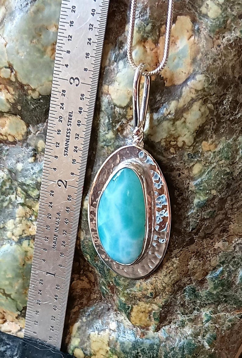 K&ouml;nnte beinhalten: Ovale Anh&auml;nger Halskette mit einem gro&szlig;en, hellblauen Edelstein, ca. 3,8 cm lang, in einem silberfarbenen Metallrahmen gefasst. Der Anh&auml;nger h&auml;ngt an einer zarten Silberkette. Der Edelstein hat ein marmoriertes Aussehen. Der Hintergrund ist eine gesprenkelte gr&uuml;n-braune Oberfl&auml;che.