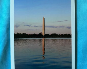 May include: A postcard featuring the Washington Monument reflected in the calm water of the Tidal Basin. The sky is a gradient of blue and the monument is a light beige color. The water reflects the monument and sky.