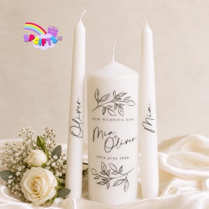 May include: A set of three white candles comprising one thick pillar candle and two slender taper candles. The pillar candle features black text reading Our Wedding Day, Mia + Oliver, and 19th June 2020 with leaf illustrations. The tapers are personalised with the names Oliver and Mia.