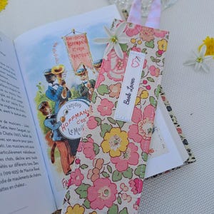 Peut inclure: Marque-page en tissu fleuri avec une poignée rayée rose et blanche, inséré dans un livre ouvert. Le marque-page porte une étiquette blanche avec l'inscription "Book Lover". Le tissu présente des fleurs roses, jaunes et vertes.