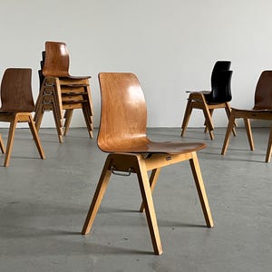 May include: A collection of wooden chairs with a mid-century modern design. The chairs have a curved backrest and angled legs. Some chairs are stacked, while others stand alone. The wood has a warm, natural finish, and one chair is black.
