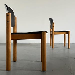 May include: Two mid-century modern chairs. The chairs have a black seat and backrest, with a curved back. The legs and frame are made of light-colored wood. The chairs are set against a white wall.