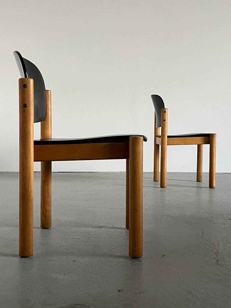 May include: Two mid-century modern chairs. The chairs have a black seat and backrest, with a curved back. The legs and frame are made of light-colored wood. The chairs are set against a white wall.
