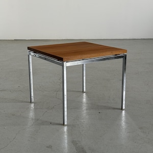 Op de afbeelding: Een vierkante bijzettafel met een houten blad en een verchroomd metalen frame. De tafel heeft een minimalistisch ontwerp, met vier rechte poten. Het tafelblad is warmbruin. Afmetingen: ongeveer 45 cm x 45 cm.