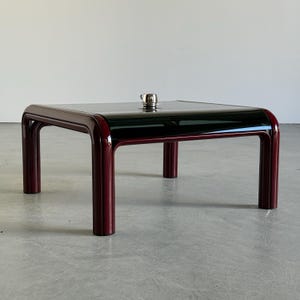 Op de afbeelding: Een vierkante salontafel met een zwarte glazen bovenkant en bordeauxrode afgeronde poten. Een klein, zilverkleurig object staat op het tafeloppervlak. De tafel heeft een modern, minimalistisch ontwerp.