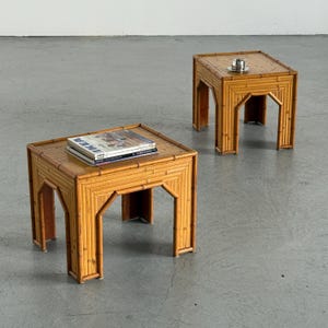 May include: Two square side tables constructed from light brown bamboo stalks. Each table features a flat top surface and four legs with arched cutouts. One table holds a stack of magazines, while the other holds a small silver metal cup.