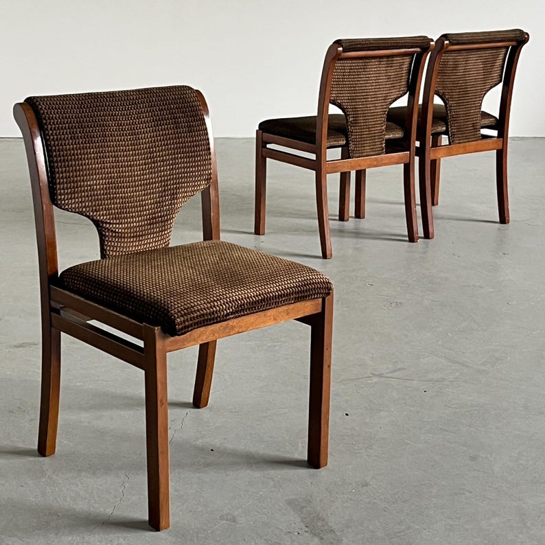 May include: Four wooden chairs with brown patterned fabric seats and backs. The chairs have a mid-century modern design, with a curved back and straight legs. The fabric has a textured appearance, and the wood has a warm, natural finish.