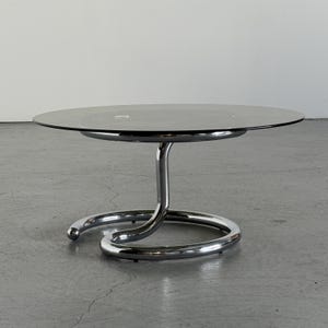 以下が含まれることがあります： スモークグレーの円形ガラス天板を備えたコーヒーテーブル。クロームメッキされた金属製のベースは、S字型にカーブした支柱が床面の広い円形リングへと繋がる、一体型の構造で天板を支えています。