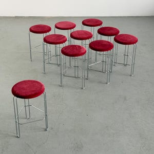May include: A collection of ten bar stools with a minimalist design. Each stool features a round, crimson red velvet seat atop a slender, chrome-finished metal frame. The stools are arranged on a gray concrete floor.