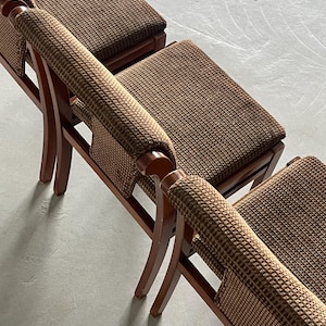 May include: Three brown wooden chairs with woven brown fabric seats and armrests. The chairs have a curved design and are arranged in a row, showcasing a classic, vintage aesthetic. The chairs are set against a neutral gray background.
