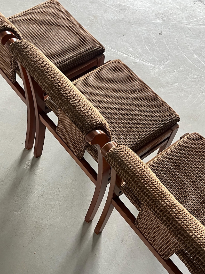 May include: Three brown wooden chairs with woven brown fabric seats and armrests. The chairs have a curved design and are arranged in a row, showcasing a classic, vintage aesthetic. The chairs are set against a neutral gray background.