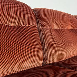 May include: Close-up view of a section of a burnt orange velvet sofa. The fabric has a subtle checkered pattern in darker orange and black. The sofa features a tufted design with distinct button tufting on the cushions. The image highlights the texture and color of the upholstery, showcasing the quality and detail of the sofa's construction. The style is mid-century modern.