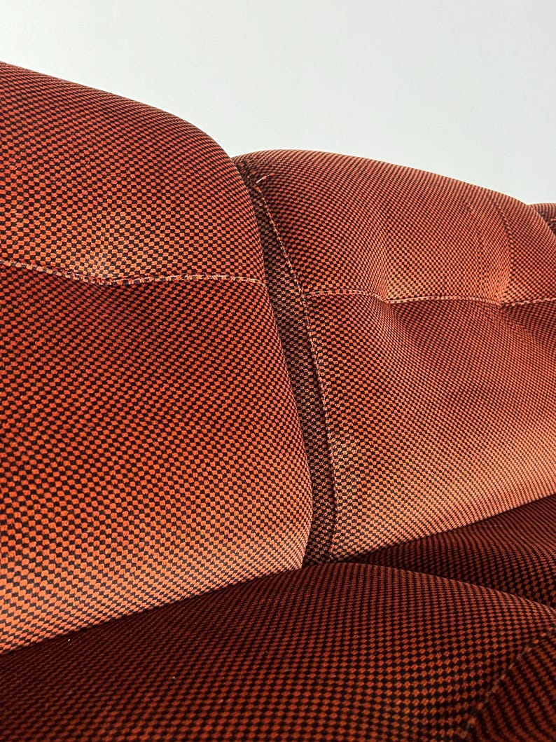 May include: Close-up view of a section of a burnt orange velvet sofa. The fabric has a subtle checkered pattern in darker orange and black. The sofa features a tufted design with distinct button tufting on the cushions. The image highlights the texture and color of the upholstery, showcasing the quality and detail of the sofa's construction. The style is mid-century modern.