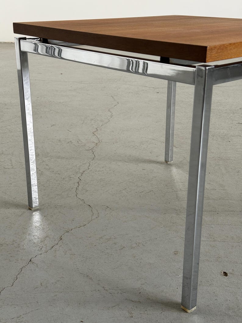 Op de afbeelding: Een vierkante salontafel in mid-century modern stijl. Het tafelblad is warmbruin en de poten en het frame zijn verchroomd. De tafel heeft een minimalistisch ontwerp, met strakke lijnen en een eenvoudig silhouet. De tafel is ongeveer 61 cm lang, 61 cm breed en 46 cm hoog.