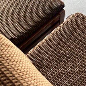 May include: Close-up of two brown woven fabric chairs with wooden frames. The fabric has a textured, grid-like pattern. The chairs are angled, showing the seat and part of the backrest. The color is a warm, earthy brown.