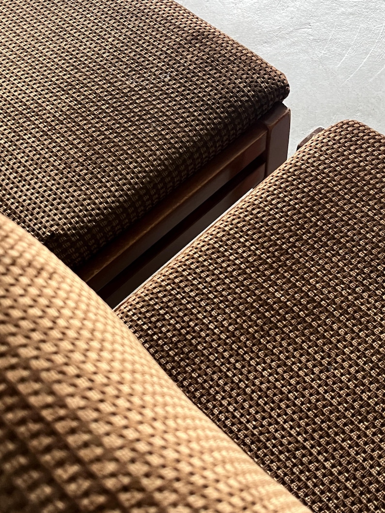May include: Close-up of two brown woven fabric chairs with wooden frames. The fabric has a textured, grid-like pattern. The chairs are angled, showing the seat and part of the backrest. The color is a warm, earthy brown.