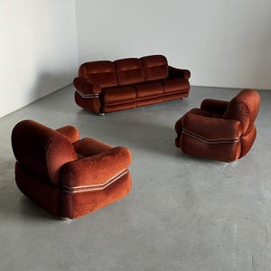 May include: A three-piece vintage living room set in a rich burnt orange corduroy upholstery. The set includes a three-seater sofa and two armchairs. Each piece features clean lines and minimalist design with subtle metallic trim accents. The furniture exudes a retro 70s aesthetic, ideal for mid-century modern or contemporary home decor.