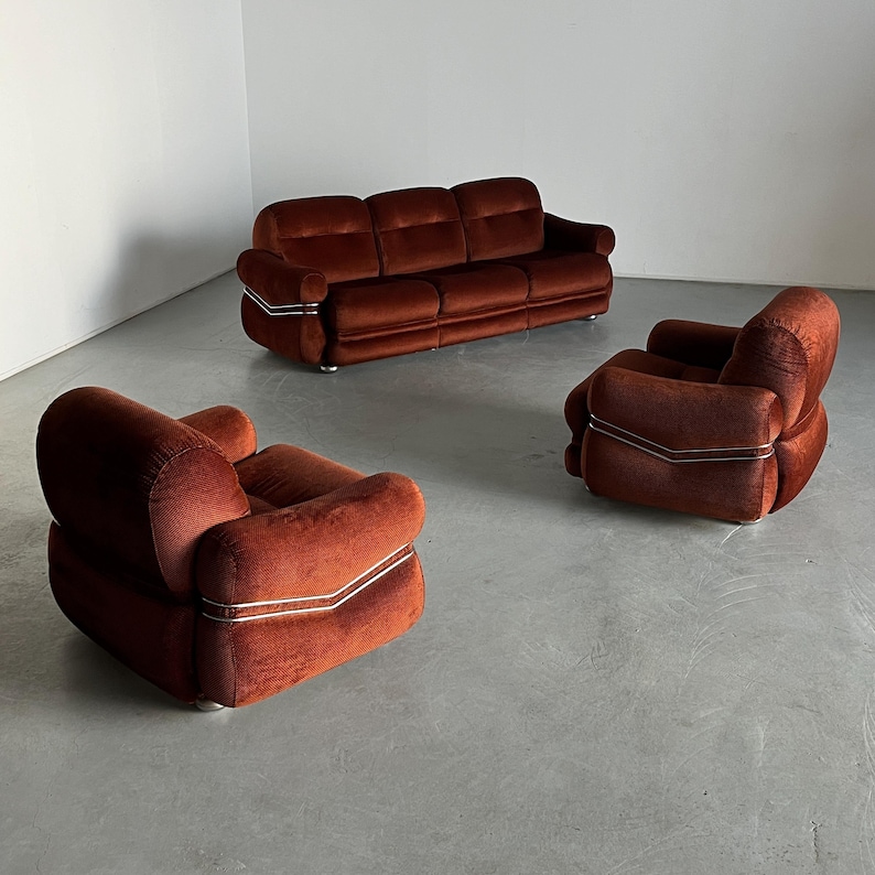 May include: A three-piece vintage living room set in a rich burnt orange corduroy upholstery. The set includes a three-seater sofa and two armchairs. Each piece features clean lines and minimalist design with subtle metallic trim accents. The furniture exudes a retro 70s aesthetic, ideal for mid-century modern or contemporary home decor.