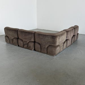 May include: A large, brown sectional sofa with a geometric pattern. The sofa has a low profile and a curved design. The base is trimmed with a silver metal. The sofa is placed against a white wall.