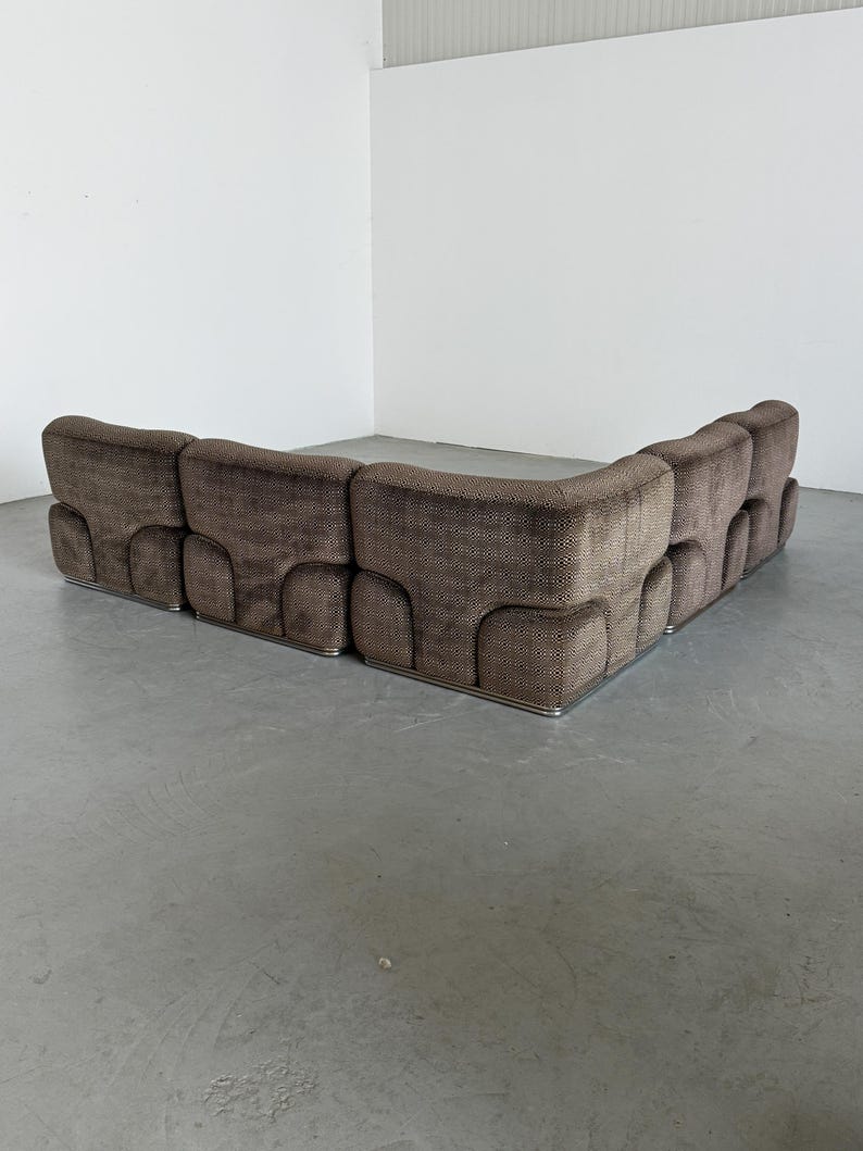 May include: A large, brown sectional sofa with a geometric pattern. The sofa has a low profile and a curved design. The base is trimmed with a silver metal. The sofa is placed against a white wall.