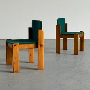 Op de afbeelding: Twee houten stoelen met donkergroene zitkussens en rugleuningen. De stoelen hebben een eenvoudig, geometrisch ontwerp met vierkante poten en een natuurlijke houten afwerking. De stoelen staan op een grijze ondergrond.