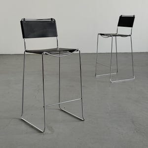 Puede incluir: Dos taburetes de bar modernos con asientos y respaldos de cuero negro. Los taburetes tienen un diseño minimalista con una estructura de metal cromado. Los asientos son rectangulares y los respaldos cuadrados. Están sobre un suelo de hormigón gris.