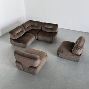 May include: A brown modular sofa set with a corner sectional and two armchairs. The furniture has a textured fabric and a low profile design. The set is placed on a gray floor, creating a modern and stylish living room arrangement.