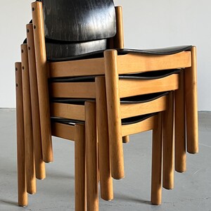 May include: A stack of mid-century modern chairs. The chairs have black seats and backs, with a curved design. The frames and legs are made of light-colored wood. The chairs are stacked on top of each other.
