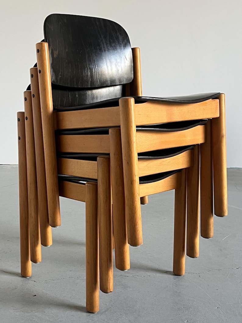 May include: A stack of mid-century modern chairs. The chairs have black seats and backs, with a curved design. The frames and legs are made of light-colored wood. The chairs are stacked on top of each other.