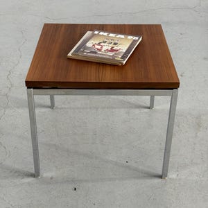 Op de afbeelding: Een vierkante salontafel in mid-century modern stijl met een donkerhouten blad en chromen poten. Op de tafel ligt een tijdschrift met de tekst "IKEA 98". De tafel is ongeveer 46 cm hoog en 76 cm breed.