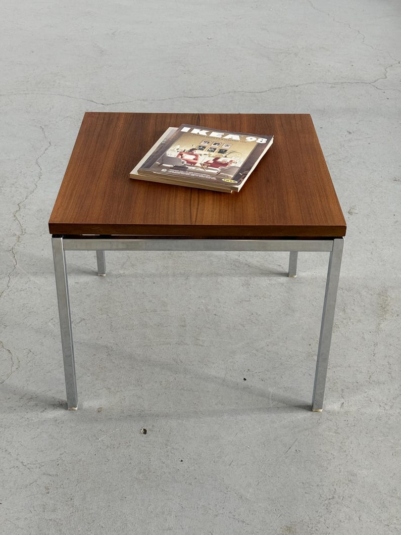 Op de afbeelding: Een vierkante salontafel in mid-century modern stijl met een donkerhouten blad en chromen poten. Op de tafel ligt een tijdschrift met de tekst "IKEA 98". De tafel is ongeveer 46 cm hoog en 76 cm breed.