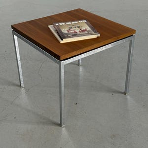 Op de afbeelding: Vierkante bijzettafel met een donkerhouten blad en een zilverkleurig metalen frame. De tafel heeft vier rechte poten. Twee boeken liggen op het tafelblad, met de tekst "IKEA 98" zichtbaar op het bovenste boek.