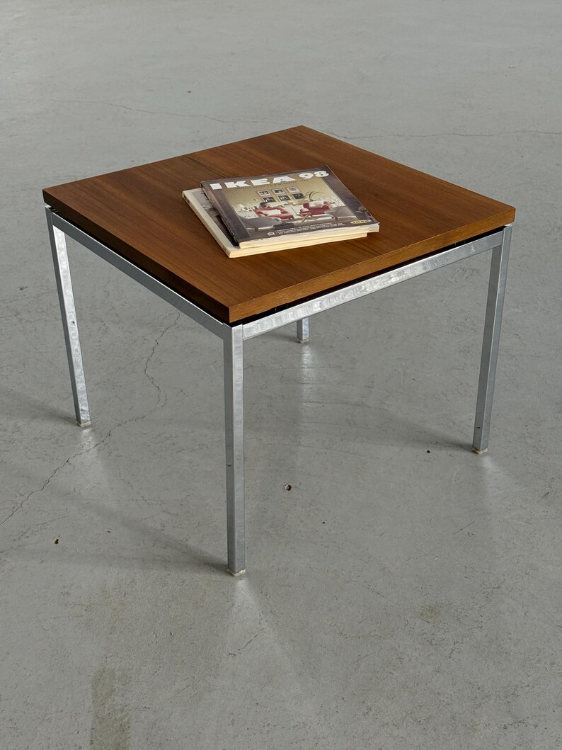 Op de afbeelding: Vierkante bijzettafel met een donkerhouten blad en een zilverkleurig metalen frame. De tafel heeft vier rechte poten. Twee boeken liggen op het tafelblad, met de tekst "IKEA 98" zichtbaar op het bovenste boek.