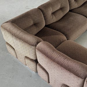 May include: A brown sectional sofa with a geometric pattern. The sofa has a low profile and a modular design, with multiple sections arranged in an L-shape. The fabric has a repeating pattern of small squares.