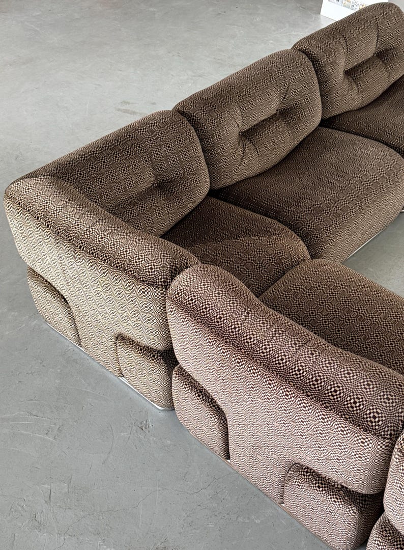 May include: A brown sectional sofa with a geometric pattern. The sofa has a low profile and a modular design, with multiple sections arranged in an L-shape. The fabric has a repeating pattern of small squares.