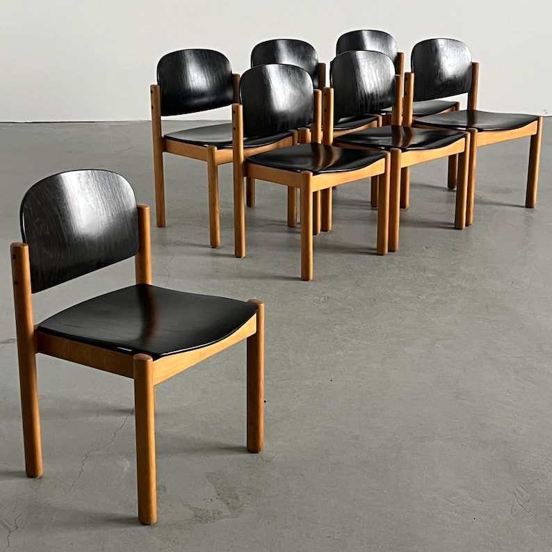 May include: A set of eight wooden chairs with black seats and backs. The chairs have a simple, minimalist design with light-colored wooden frames and dark, curved backs. The chairs are arranged in a group, suggesting they are for a dining or seating area.