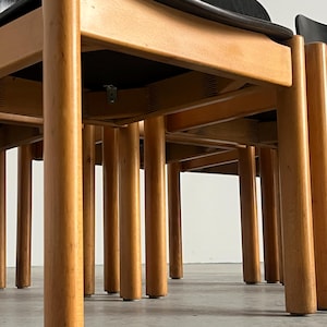 May include: A set of wooden chairs with black seats and backs. The chairs have light brown, cylindrical legs and a natural wood finish. The design features a minimalist aesthetic, with clean lines and a focus on the wood's grain.