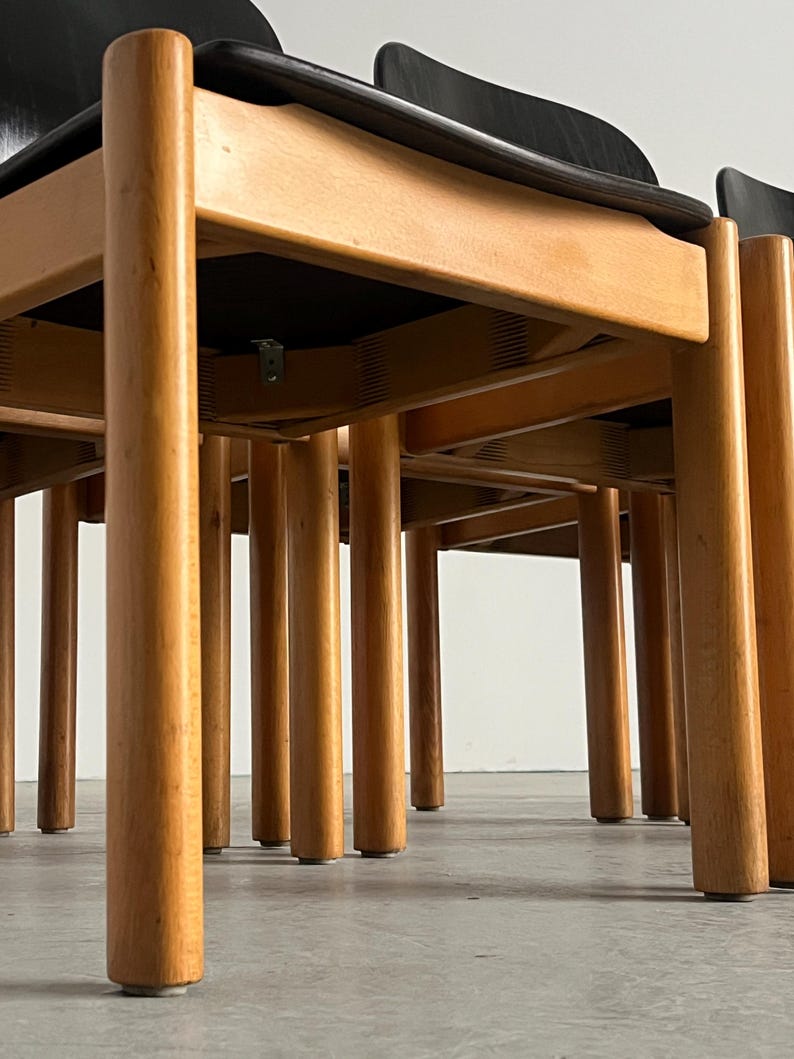 May include: A set of wooden chairs with black seats and backs. The chairs have light brown, cylindrical legs and a natural wood finish. The design features a minimalist aesthetic, with clean lines and a focus on the wood's grain.