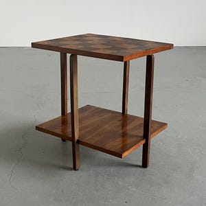 May include: A two-tiered wooden side table. The top surface features a checkerboard pattern, while the lower shelf and legs have a smooth, polished finish. The table is made of dark brown wood.