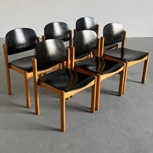 May include: A set of seven mid-century modern chairs. Each chair has a black seat and backrest, with a curved back design. The frames are made of light-colored wood, and the chairs are arranged in a group.