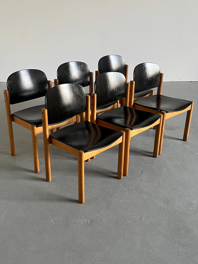 May include: A set of seven mid-century modern chairs. Each chair has a black seat and backrest, with a curved back design. The frames are made of light-colored wood, and the chairs are arranged in a group.