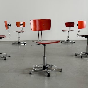 Peut inclure: Plusieurs chaises réglables rouges et chromées sont disposées dans une pièce. Les chaises ont un dossier et une assise arrondis, avec une base chromée et un repose-pieds. Elles sont placées devant un mur blanc et un sol gris.