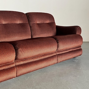 May include: A rich, dark reddish-brown corduroy sofa with a tufted design. The sofa features a three-cushion seat and rolled arms.  The fabric has a subtle textured pattern. The sofa is positioned on a light gray floor, showcasing its low profile and clean lines. This vintage-style sofa is ideal for mid-century modern or retro-inspired interiors.