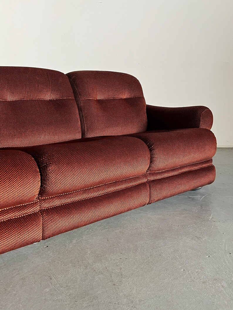 May include: A rich, dark reddish-brown corduroy sofa with a tufted design. The sofa features a three-cushion seat and rolled arms.  The fabric has a subtle textured pattern. The sofa is positioned on a light gray floor, showcasing its low profile and clean lines. This vintage-style sofa is ideal for mid-century modern or retro-inspired interiors.